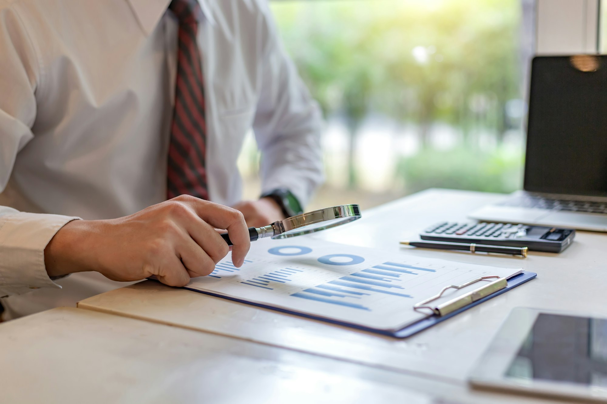 Businessman holding a magnifying glass with graph information document on the desk.